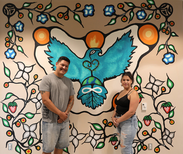 Artists Jaylene Cardinal and Dakota Ward stand in front of their completed mural, featuring an eagle, a heart-shaped tree, a sacred fire and vines of flowers, leaves and strawberries. Jaylene is wearing a black tank top and blue jeans, her dark-brown hair in a ponytail. Dakota is wearing a grey t-shirt and blue jean shorts and short dark-brown hair.