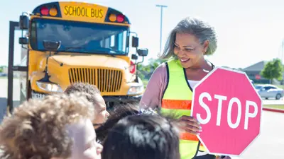 A female crossing guard holding a stop sign standing outside in front of a school bus with multiple children