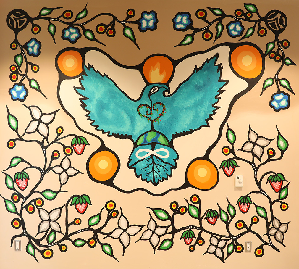 A painted mural on an interior wall of the Rideau Heights Community Centre. The mural features a blue eagle and symbols for the earth, a heart-shaped tree, an Infinity symbol and roots. Surrounding the eagle are orbs of orange symbolizing energy and sacred fire. Vines of flowers, leaves and strawberries border the mural.