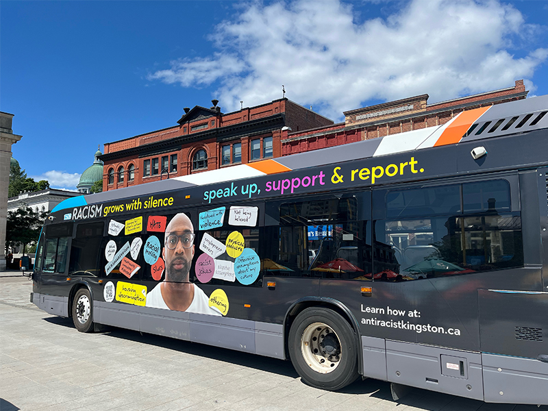Kingston Transit bus with Anti-Racist campaign wrap. Text reads, "Racism grows with silence. Speak up, support & report. Learn how at antiracistkingston.ca"