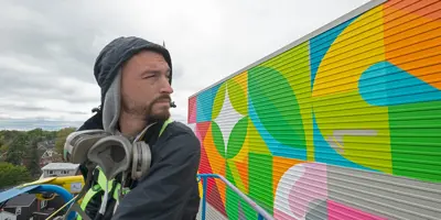 Painter painting the large mural on the side of a building while utilizing a cherry picker