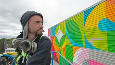 Painter painting the large mural on the side of a building while utilizing a cherry picker